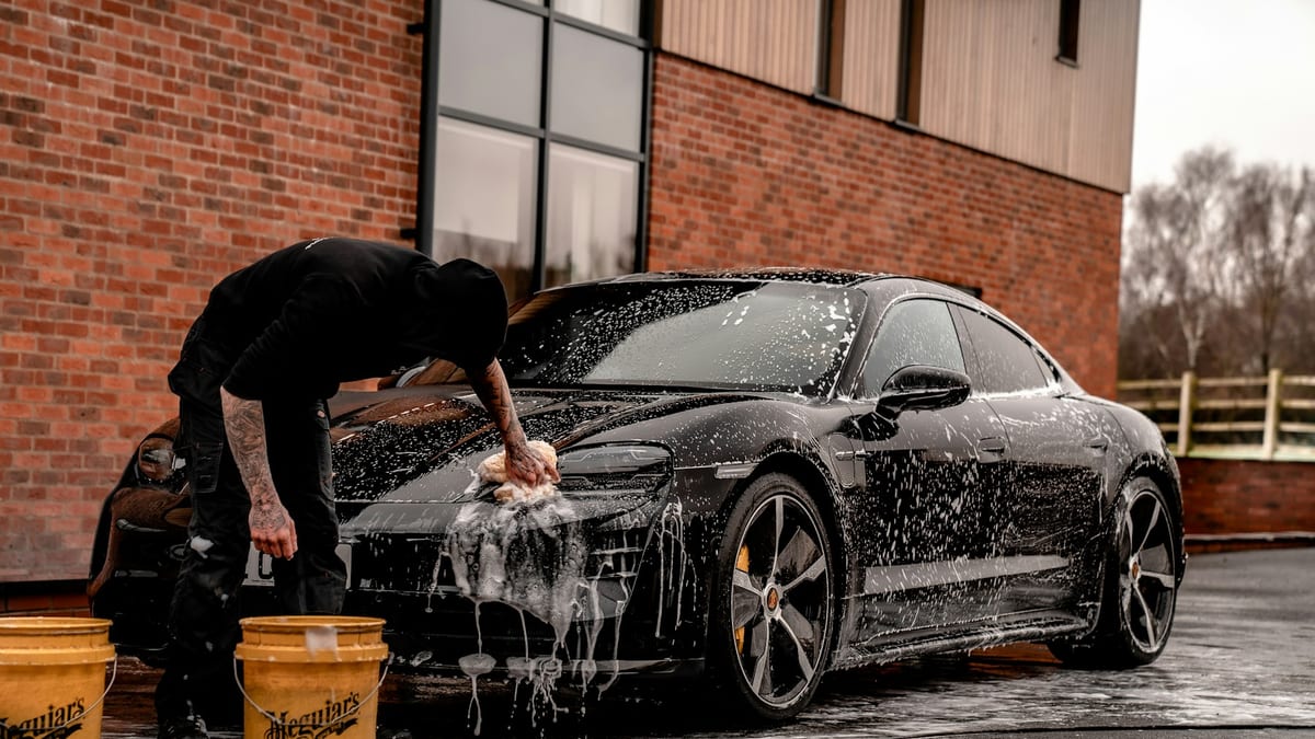 A detailer hand-washing a glossy black sports car covered in thick white foam at a professional detailing studio.
