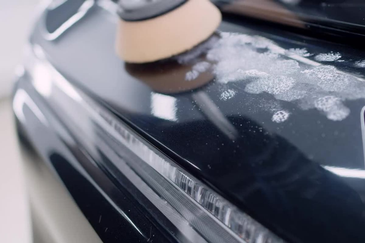 A dual-action polisher with a foam pad working the glossy black hood of a car, the headlight reflecting studio light.