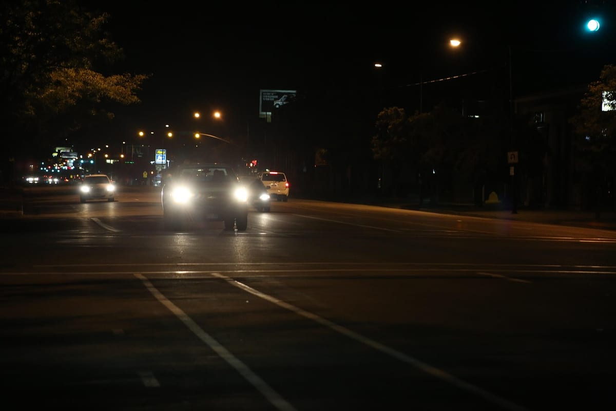 A car's bright headlight beams glaring at an intersection on a dark city street at night, with streetlights in the background.