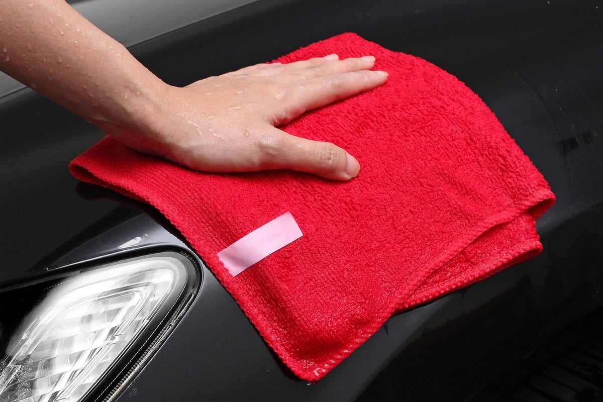 A hand pressing a red microfiber towel against a corrected black car panel next to the headlight, wiping the surface after polishing.