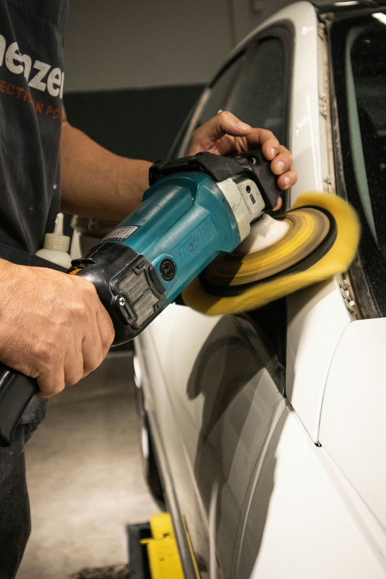 A technician running a rotary polisher with a foam pad along the rear quarter panel and window trim of a white car.