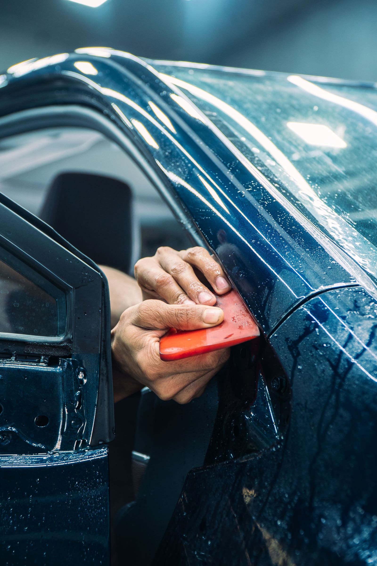 A technician hand-fitting protective film to a dark blue car door with a squeegee.