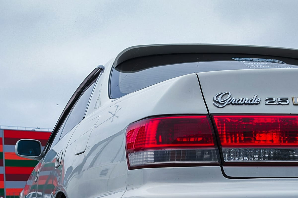Close-up of a silver sedan's dark tinted rear window and taillight at a car meet, showing a deep, uniform tint tier from the rear glass down through the C-pillar.